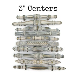 May include: A collection of ten drawer pulls with 3" centers, featuring various decorative shapes and a distressed finish in shades of cream, grey, and metallic gold. Each handle displays unique textures, including ribbed, smooth, and ornate patterns.