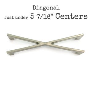 May include: A silver-toned, X-shaped cabinet pull with a diagonal design. The text above the pull reads "Diagonal Just under 5 7/16" Centers". The hardware has a modern, minimalist aesthetic.