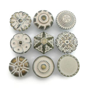 May include: Nine decorative cabinet knobs in various designs. The knobs feature a distressed white and gold finish. Styles include floral, geometric, and circular patterns. The knobs are arranged on a white background.