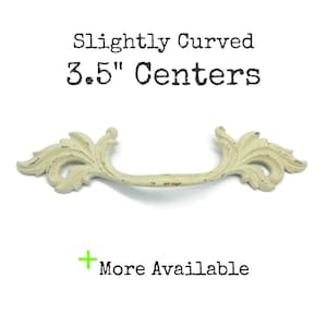 May include: A cream-colored drawer pull with a distressed finish and ornate leaf-like ends. The text above reads Slightly Curved 3.5" Centers, and the text below includes a green plus sign followed by the words More Available.