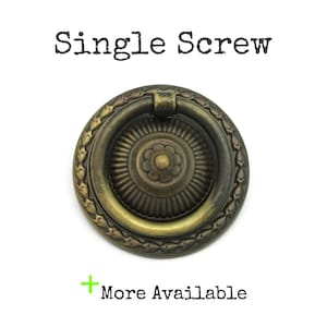 May include: Antique brass-toned drawer pull with a circular design. The pull features a central floral motif surrounded by a ring and a decorative outer edge. The text "Single Screw" is above the pull, and "More Available" is below.