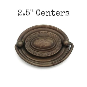 May include: Antique bronze oval drawer pull with a decorative center and a curved handle. The text "2.5" Centers" is above the pull. The pull measures 2.5 inches between the mounting holes.