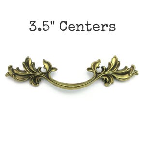 May include: An antique brass-colored drawer pull with ornate leaf and scroll detailing. The handle is 3.5 inches (8.89 cm) between the centers of the mounting holes. The text "3.5" Centers" is above the handle.