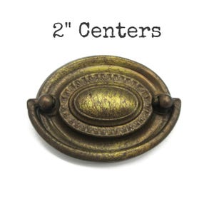 May include: An oval-shaped brass-toned metal drawer pull with a distressed finish and a swinging handle. The hardware features an ornate border and a central oval plate. The text 2" Centers is printed above the object, indicating the mounting distance.