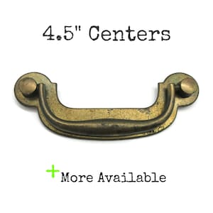 May include: A brass-toned metal drawer pull with a curved handle and two circular mounting plates. The text 4.5" Centers is printed above the handle, and the text + More Available is printed below the handle in black lettering.
