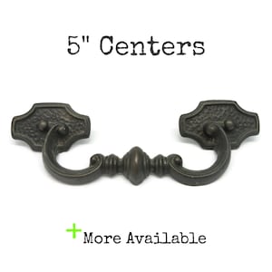 May include: A dark bronze-toned metal drawer pull with a swinging handle and two textured mounting plates. The text 5" Centers is printed above the hardware, and the text + More Available is printed below the hardware on a white background.