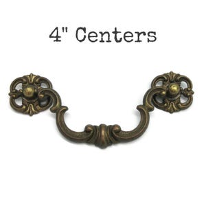 May include: An antique brass-toned metal drawer pull with a curved handle and two ornate circular mounting plates. The hardware features intricate scrollwork details and a weathered finish. The text 4" Centers is printed above the handle.