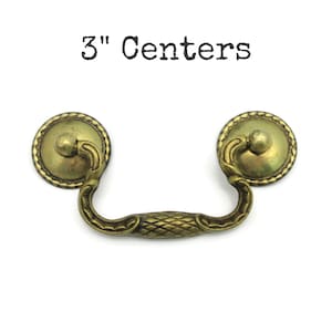 May include: Antique brass drawer pull with a decorative design. The pull has a curved handle with a diamond pattern and two round backplates. The text "3" Centers" is above the pull.