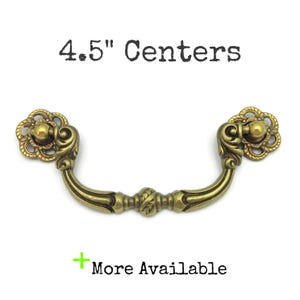 May include: Antique brass drawer pull with a floral design on the ends and a curved handle. The text "4.5" Centers" is above the pull, and "More Available" is below.