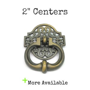 May include: An antique brass-toned metal drawer pull with a circular, ornate filigree backplate and a swinging drop handle. The text 2" Centers is printed above the hardware, and the text + More Available is printed below the hardware.
