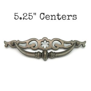 May include: Antique-style drawer pull with a decorative design. The pull is a muted silver color and features a curved handle and ornate detailing. The text "5.25" Centers" is printed above the pull.