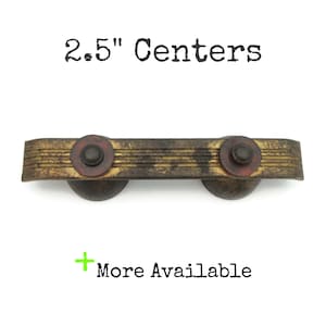 May include: A vintage-style cabinet pull with a distressed brass finish. The pull features a rectangular bar with horizontal grooves and two circular end pieces with red accents. The text "2.5" Centers" is above the pull, and "More Available" is below.