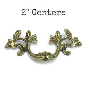 Peut inclure: Une poignée de tiroir en métal couleur laiton antique présentant un design orné et symétrique avec des motifs de feuilles. La quincaillerie a une forme incurvée et inclut le texte 2" Centers imprimé en noir au-dessus de l'objet.