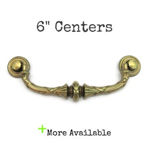 May include: Antique brass-toned drawer pull with a decorative design. The pull has a curved handle with ornate detailing and circular end pieces. The text "6" Centers" is above the pull, and "More Available" is below.