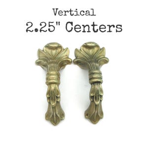 May include: Two antique brass-toned drawer pulls with a vertical design. Each pull features a floral top and a detailed stem. The text "Vertical 2.25" Centers" is printed above the pulls.