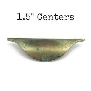 May include: A vintage-style brass drawer pull with a curved design. The pull has a distressed finish with green patina and measures 1.5 inches between the centers. The text "1.5" Centers" is printed above the pull.