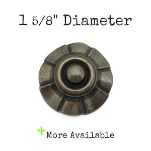 May include: A bronze-toned cabinet knob with a diameter of 1 5/8 inches. The knob has a circular design with a central raised button and a decorative, ridged outer edge. Text above reads "1 5/8" Diameter". The text "More Available" is below.