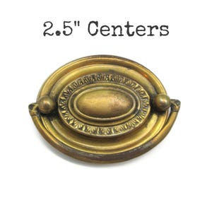 May include: An oval brass drawer pull with a central medallion and a swinging handle. The hardware features a weathered gold finish and decorative detailing around the center. The text 2.5" Centers is printed above the object.