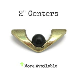May include: Gold metal drawer pull with a black round knob in the center. The pull is shaped like a V and has a 2" center measurement.