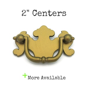 May include: A gold-colored drawer pull with a decorative design. The pull has a curved handle and is labeled "2" Centers". The text "More Available" is also visible.