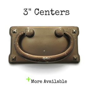 May include: A rectangular bronze-toned metal drawer pull with a swinging bail handle and four pyramid-shaped corner accents. The hardware features visible surface wear and paint chips. The text 3" Centers and + More Available is present.
