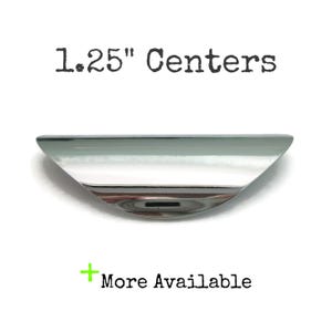 May include: A shiny, silver-colored cabinet pull with a curved design. The text "1.25" Centers" is above the pull, and "More Available" is below. The pull measures 1.25 inches in length.