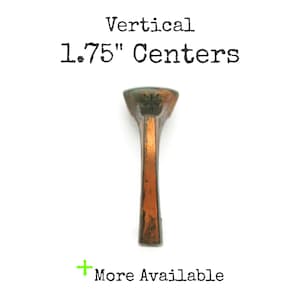 May include: A single, aged bronze-colored cabinet pull with a vertical design. The text "Vertical 1.75" Centers" is printed above the pull. The words "More Available" are printed below the pull.