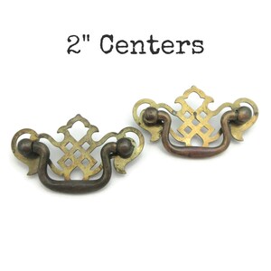 May include: Two antique brass drawer pulls with a decorative lattice design. The pulls have a dark bronze finish on the handles and measure 2 inches between the centers. The text "2" Centers" is above the pulls.