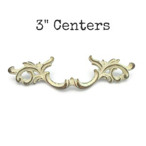 May include: Off-white and gold decorative drawer pull with a floral design. The text "3" Centers" is above the drawer pull. The drawer pull measures 3 inches between the screw holes.