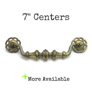 May include: A brass-toned metal drawer pull with a central spindle design and two circular, scalloped mounting plates. The text 7" Centers is printed above the handle, and the text + More Available is printed below the handle.