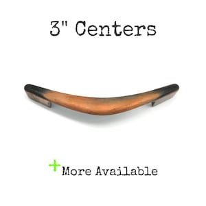 May include: A curved wooden cabinet pull with a 3-inch center-to-center measurement. The pull is a warm brown color with dark accents on the ends. The text "3" Centers" is above the pull, and "More Available" is below.