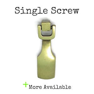 May include: A brass-coloured metal drop pull handle featuring a rectangular mounting block at the top and a smooth, flat, spade-shaped pendant. The text "Single Screw" appears above the object, and "+ More Available" is printed below.