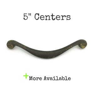 May include: Antique bronze-colored cabinet pull with a curved design. The text "5" Centers" is above the pull, and "More Available" is below. The pull measures 5 inches between the centers of the mounting holes.