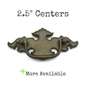 May include: Antique brass drawer pull with a decorative design. The pull has a curved handle and measures 2.5 inches between the centers of the mounting holes. The text "2.5" Centers" is above the pull, and "More Available" is below.