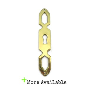 May include: A brass-colored keyhole cover with an elongated, symmetrical design. The cover features a keyhole in the center and decorative geometric shapes at the top and bottom. The text "More Available" is at the bottom.