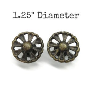 May include: Two round metal knobs with an antique bronze finish, featuring a cutout petal design radiating from a central dome. The knobs have a 1.25" diameter as indicated by the text above them. These hardware pieces have a textured surface.