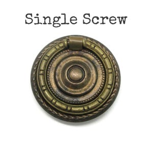 May include: A round, antique brass-toned metal drawer pull with a central circular knob and a swinging ring handle. The hardware features a decorative rope-style border and concentric circles. The text "Single Screw" is printed above the object.