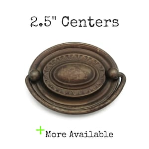 May include: Antique bronze-toned drawer pull with an oval center and a curved handle. The pull has a decorative design around the oval center and measures 2.5 inches between the centers of the mounting holes. The text "2.5" Centers" is above the pull.