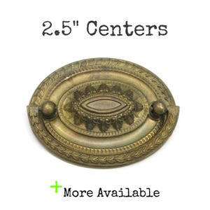 May include: Antique brass oval drawer pull with a decorative leaf and floral design. The pull has a 2.5-inch center-to-center measurement. The text "2.5" Centers" is above the pull, and "More Available" is below.