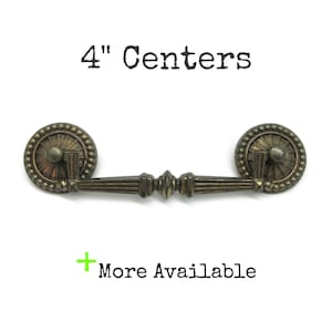 May include: A bronze-toned metal drawer pull with a central horizontal bar and two circular, beaded mounting plates. The text 4" Centers is printed above the handle, and the text + More Available is printed below the handle.
