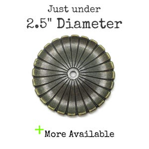 Peut inclure: Un disque métallique rond de couleur laiton antique avec un motif cannelé en forme de rayons de soleil partant d'un trou central. Le texte au-dessus indique Just under 2.5" Diameter et le texte en dessous + More Available. Finition patinée.
