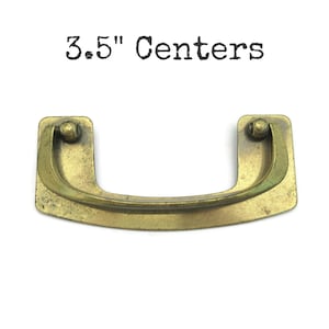 May include: A vintage-style brass drawer pull with a curved handle and rectangular mounting plates. The text "3.5" Centers" is printed above the pull. The pull measures 3.5 inches (8.89 cm) between the mounting holes.