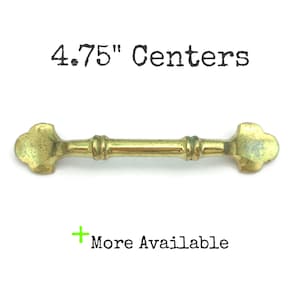 May include: Gold metal cabinet pull with a 4.75" center to center measurement. The pull has a simple, elegant design with a slightly curved shape and a decorative detail in the center.