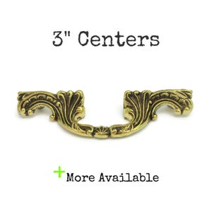 May include: A gold-toned metal cabinet pull with a decorative, ornate design. The pull is 7.6 cm between the mounting holes.