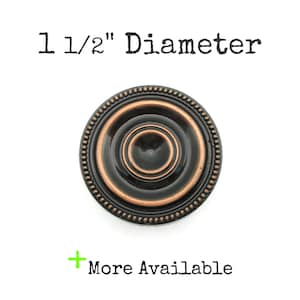 May include: A round, bronze-colored cabinet knob with a diameter of 1 1/2 inches. The knob features a beaded outer edge and concentric circles, with the text "1 1/2" Diameter" above and "More Available" below.