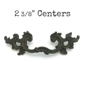 May include: Ornate bronze-toned drawer pull with a decorative scroll design. The pull has a center-to-center measurement of 2 3/8 inches. The text "2 3/8" Centers" is printed above the pull.