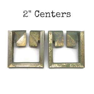 May include: Two square-shaped metal drawer pulls with a weathered, tarnished brass finish and a geometric cutout design. The text 2" Centers is printed above the hardware, indicating the distance between the mounting points on each pull.