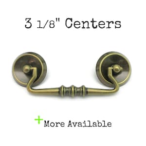May include: Antique brass drawer pull with a curved handle and circular backplates. The text "3 1/8" Centers" is printed above the pull. The text "More Available" is printed below the pull.