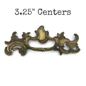 May include: Ornate antique brass drawer pull with a decorative design. The pull measures 3.25 inches between the centers of the mounting holes. The text "3.25" Centers" is printed above the pull.