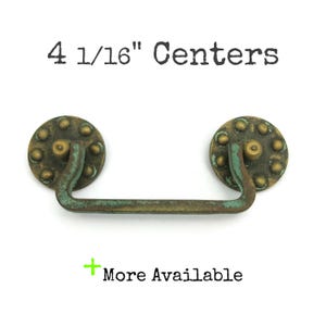 May include: A brass cabinet pull with a distressed green finish and a 4 1/16" centre to centre measurement. The pull has a round, decorative plate with raised dots.